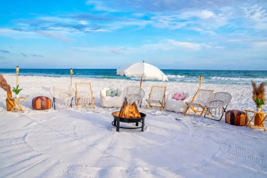 A luxury beachside bonfire in Santa Rosa Beach FL along 30A with a fringe umbrella, white lounge chairs, bean bags, and leather poufs around a lit fire pit.