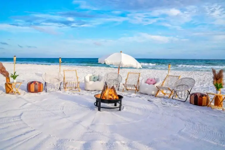 A luxury beachside bonfire in Santa Rosa Beach FL along 30A with a fringe umbrella, white lounge chairs, bean bags, and leather poufs around a lit fire pit.