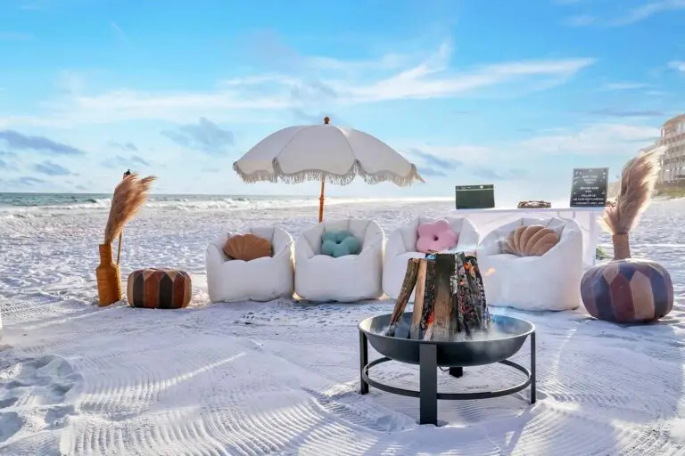 A luxury beach bonfire setup on 30A near Destin, Florida, featuring white bean bags, flower pillows, and a fringe beach umbrella.